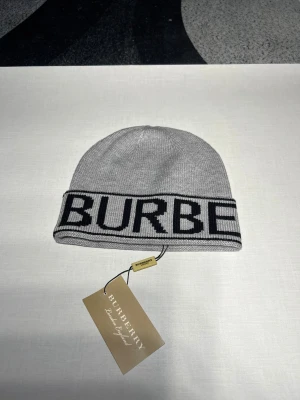 Grå Burberry mössa med logotyp - Snygg grå mössa från Burberry med bred uppvikt kant och stor svart logotyp runt om. Mössan är stickad i mjukt material och har en klassisk, rund form. Perfekt för dig som vill ha en clean look med tydlig designerstil.