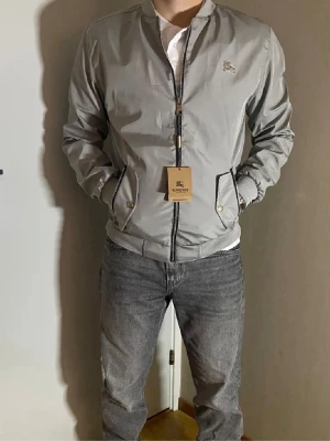 Burberry Bomberjacka - Snygg grå bomberjacka från Burberry med ribbade muddar, dragkedja framtill och två fickor med både dragkedja och knapp. Jackan har en minimalistisk look med Burberry-logga på bröstet och är tillverkad i ett lätt, syntetiskt material.