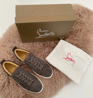 Grå mocka sneakers Christian Louboutin - Snygga grå sneakers från Christian Louboutin i mjuk mocka med vita platta sulor och ikonisk röd undersida. Skorna har rund tå, snörning och beige innersida. Perfekt för dig som vill ha en stilren och lyxig look med exklusiv känsla.