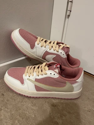 Nike Air Jordan 1 Low rosa/vit - Nike Air Jordan 1 Low sneakers i en snygg kombo av ljusrosa och vitt med beige snörning. Ovandelen är i mocka och skinn, med klassisk rund tå och platt sula. Yttersulan är rosa och loggan syns på tungan och hälen. Perfekt för dig som gillar stilrena och trendiga sneakers.