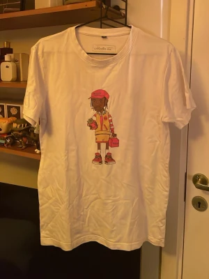 Vit t-shirt Mister Tee med grafiskt tryck - Vit t-shirt från Mister Tee i storlek S med ett färgstarkt grafiskt tryck på framsidan som visar en tecknad figur med röda och gula detaljer. Klassisk rund hals och korta ärmar. Materialet är mjuk bomull som känns skönt mot huden.