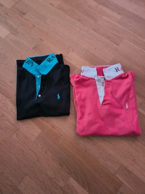 Ralph Lauren pikétröjor svart & rosa - Två klassiska pikétröjor från Ralph Lauren. En svart med turkos krage och logga, storlek M. En rosa med vit krage och logga, storlek L. Båda har tryck på kragen och knappar framtill. Perfekta för en sportig och stilren look.