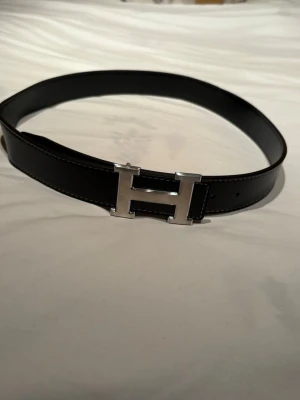Svart Her-mès skärp med H-spänne - Säljer ett svart Her-mès skärp med ikoniskt H-spänne i silverfärgad metall. Bältet är gjort av äkta skinn med tydliga vita sömmar längs kanterna och flera hål för justering. Perfekt accessoar för att lyfta din outfit.