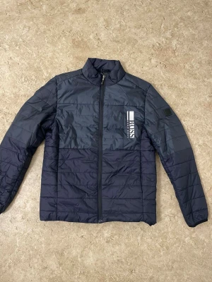 Mörkblå quiltad jacka från Boss - Snygg mörkblå quiltad jacka från Boss med diskret logga på bröstet. Jackan har ståkrage, hel dragkedja och är tillverkad i lätt polyester. Perfekt för kyliga dagar och har en stilren look med horisontella sömmar.