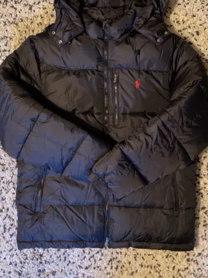 Svart dunjacka från Ralph Lauren - Svart dunjacka från Ralph Lauren med röd logga på bröstet. Jackan har huva, flera dragkedjeförsedda fickor och RL-detalj på dragkedjan. Puffig modell som håller värmen, perfekt för kyliga dagar. Klassisk och stilren design.