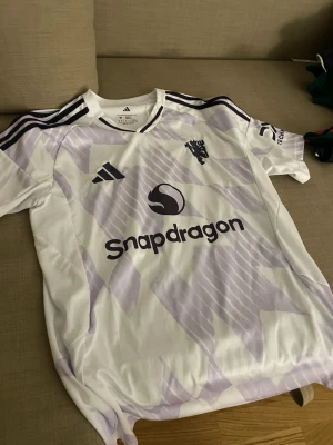 Vit/lila Adidas fotbollströja M - Snygg fotbollströja från Adidas i slim fit, storlek M. Tröjan är vit med ljuslila geometriskt mönster och svarta detaljer, inklusive Adidas-logga och Manchester United-emblem. Materialet är lätt polyester och har svarta ränder på axlarna.
