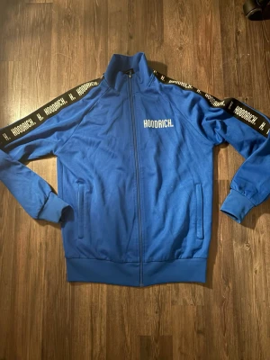 Blå track jacket från Hoodrich - Säljer en blå track jacket från Hoodrich med vit logga på bröstet och svarta band med vit text längs axlarna. Jackan har hel dragkedja, ståkrage och två fickor framtill. Perfekt för en sportig och street-inspirerad look.