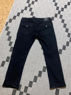 Svarta True Religion Ricky jeans - Svarta jeans från True Religion, modell Ricky Relaxed Straight. Klassisk femficksdesign med snygga bakfickor och silverfärgade knappar. Raka ben och normal passform, tillverkade i slitstarkt bomullsmaterial. Perfekt för dig som gillar streetwear och vill ha en clean look.
