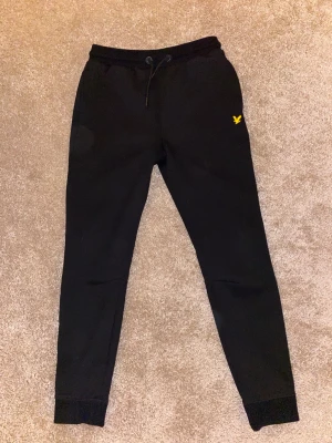 Svarta mjukisbyxor från Lyle & Scott - Svarta mjukisbyxor från Lyle & Scott med gul broderad logga på vänster lår. Byxorna har snörning i midjan och ribbade muddar vid bensluten. Sköna och stilrena, perfekta för chill eller träning.