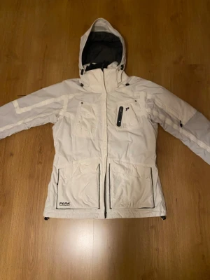 Vit vinterjacka från Peak Performance - Snygg vit vinterjacka från Peak Performance med huva, flera praktiska fickor och dragkedja framtill. Jackan har en sportig look och är perfekt för kalla dagar. Stora fickor framtill och diskreta svarta detaljer ger en clean stil.