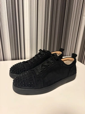 Christian Louboutin svarta sneakers med nitar - Svarta sneakers i mocka från Christian Louboutin med coola svarta nitar på tån och klassisk röd sula. Skorna har snörning och en platt sula, samt rund tå. Insidan är beige och de har en lyxig vibe som sticker ut. Storlek 43, använda lite, men inga större skador eller sådant. Inga defekter! 