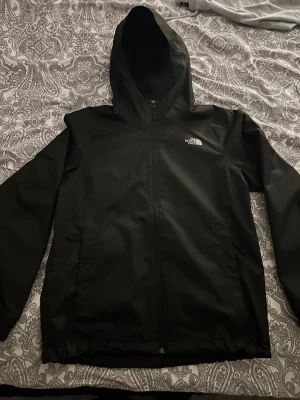 Svart vindjacka från The North Face - Svart vindjacka från The North Face med huva och hel dragkedja framtill. Jackan har två sidofickor och meshfoder på insidan för extra ventilation. Perfekt för blåsiga dagar och har en stilren look med logga på bröstet.