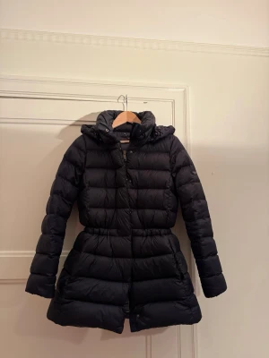 Svart dunjacka från Polo Ralph Lauren - Snygg svart dunjacka från Polo Ralph Lauren med huva och markerad midja. Jackan har långa ärmar, dragkedja med Polo-detalj och logga på ärmen. 