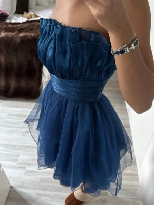 Marinblå axelbandslös kortklänning - Supersöt marinblå kortklänning med voluminös kjol i flera lager tyll och markerad midja. Axelbandslös modell med rynkad överdel och vågig kant upptill. Sitter som en xs/s .