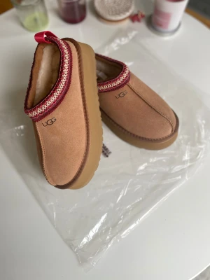 UGG slip-ins i beige mocka - UGG slip-ins i beige mocka med fluffigt foder och grov sula. Skorna har en snygg broderad kant i rött och beige samt rund tå. Perfekta för kalla dagar när du vill vara både varm och trendig.