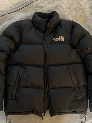 Svart pufferjacka från The North Face - Säljer en svart pufferjacka från The North Face med hög krage och dragkedja framtill. Jackan har två sidofickor med dragkedja och logga på bröstet. Perfekt för kalla dagar och riktigt snygg streetstil. Köpt för 3700kr!!