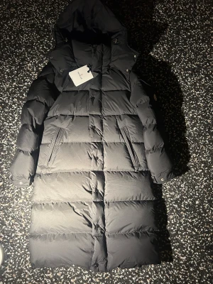 Svart lång dunjacka från Moncler - Säljer en svart, lång dunjacka från Moncler med huva och quiltad design. Jackan har Moncler-logga på ärmen, dragkedjor vid fickorna och tryckknappar framtill. Perfekt för kalla dagar med sin fluffiga och stilrena look. 