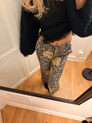 Blommiga byxor Morris & Co x H&M - Säljer ett mönstringa byxor från Morris & Co x H&M i storlek 34. De har ett blommigt mönster i beige, blått och grått med hög midja och raka ben. Materialet är mjukt och skönt, perfekt för dig som gillar statement-plagg.