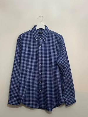 Blårutig skjorta från Polo Ralph Lauren - Klassisk blårutig skjorta från Polo Ralph Lauren med button-down krage och vita knappar. Skjortan har lång ärm och är tillverkad i mjuk bomull. Perfekt för dig som gillar en tidlös och clean stil.