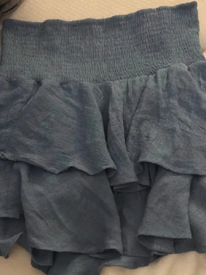 Blå volangkjol med resårmidja - Supersöt blå kortkjol med bred resår i midjan och flera lager volanger. Kjolen har en luftig och lätt känsla, perfekt för varma dagar. Materialet känns mjukt och flowy, vilket ger en avslappnad men ändå stylish vibe.