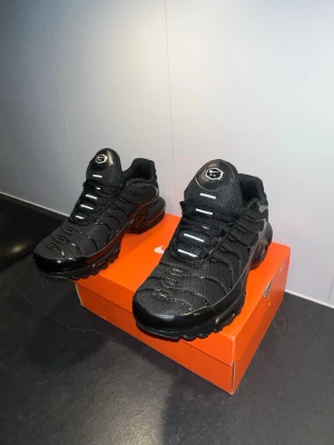 Nike skor  - Nike Air Max Plus i helsvart färg med mesh och syntetiska detaljer. Skorna har den ikoniska Tn-loggan på hälen, vågiga linjer på ovandelen och synlig Air-dämpning i sulan. Klassisk snörning och robust platt sula för extra komfort.
