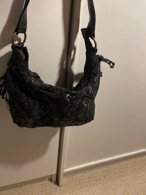 Svart axelväska med paljetter och snörning - Cool svart axelväska med broderade blommor och paljettdetaljer. Väskan har snörning upptill med metallringar och flätade band, samt dekorativa tofsar på sidorna. Perfekt för dig som gillar unika accessoarer med lite extra bling.