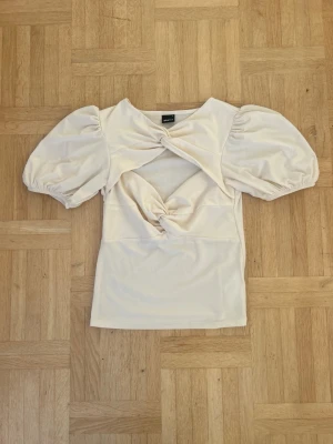 Krämvit topp med puffärm och twist💖 - Säljer en krämvit topp från Gina Tricot med puffiga korta ärmar och snygga twistade detaljer framtill. Toppen har en cut-out över magen och är gjord i mjukt stretchigt material. Perfekt för dig som vill ha något trendigt och unikt.