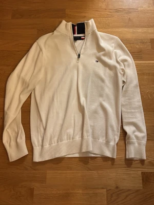 Vit half zip tröja Tommy Hilfiger - Klassisk vit långärmad tröja från Tommy Hilfiger med half zip och liten broderad logga på bröstet. Ribbad mudd vid ärmslut och nederkant samt snygg detalj med röd och marinblå rand i kragen. Perfekt till chill eller när du vill vara lite extra clean.