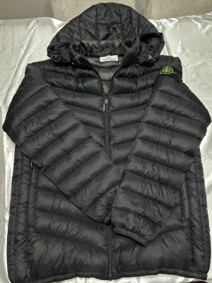 Svart pufferjacka från Stone Island - Säljer en svart pufferjacka från Stone Island med huva och dragkedja. Jackan har det klassiska Stone Island-märket på ärmen och är quiltad för extra värme. Tillverkad i polyester och har en clean, modern look som passar perfekt till höst och vinter.