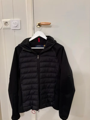 Svart pufferjacka från Polo Ralph Lauren - Svart pufferjacka från Polo Ralph Lauren med huva och dragkedja framtill. Jackan har quiltad front, släta ärmar och diskret broderad logga på bröstet. Materialet är polyester och jackan har en sportig, clean look som funkar till allt. Den enda lilla defekten är en pytte liten slitning där fram annars är allt perfekt
