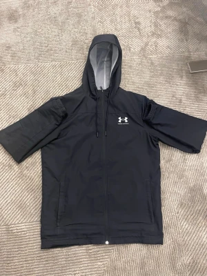 Svart vindjacka från Under Armour - Svart vindjacka från Under Armour med huva och dragkedja framtill. Jackan har en enkel design med logga på bröstet, två fickor och justerbar huva med snörning. Tillverkad i lätt syntetmaterial, perfekt för blåsiga dagar. Som passar folk med storlek s/xs