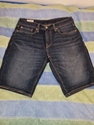 Levi's mörkblå jeansshorts - Säljer ett par klassiska mörkblå jeansshorts från Levi's med fem fickor, kontrastsömmar och knappgylf. Shortsen har normal passform och är perfekta för sommaren. Ikonisk läderpatch bak och Levi's röda etikett på bakfickan. I storlek w 28