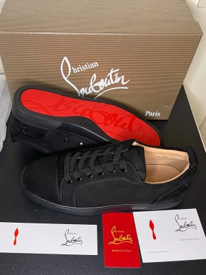 Svarta sneakers från Christian Louboutin - Säljer ett par svarta sneakers från Christian Louboutin i mocka med klassisk röd sula. Skorna har rund tå, snörning och diskreta detaljer. Insidan är beige och märkt med guldfärgad logga. Perfekta för dig som vill ha något stilrent men ändå lyxigt.