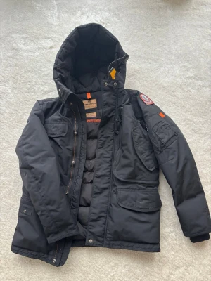 Svart Parajumpers dunjacka med huva - Säljer en svart Parajumpers dunjacka med stor huva, flera fickor och snygga orange detaljer. Jackan har patch på ärmen, dragkedja och tryckknappar framtill samt quiltad insida. Mycket bra skick/ som ny. Köpte den på kids brand store i Täby Centrum för några år sedan för 6990 kr och säljer nu för 1000 kr vid snabb affär. Stl Young medium. 