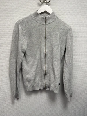 Grå kofta från Jack & Jones XS - Ljusgrå kofta från Jack & Jones i storlek XS. Tröjan har hel dragkedja framtill, ribbad krage och muddar. Tillverkad i mjukt material som passar perfekt till lager på lager under höst och vinter. Enkel och clean design utan mönster. Skriv för frågor!