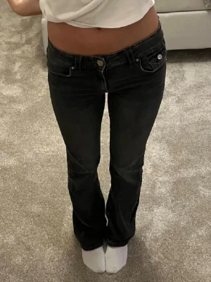 Svarta bootcut jeans med låg midja - Säljer ett par svarta jeans med låg midja och bootcut-ben. Jeansen har klassisk femficksdesign, knappgylf och snygga detaljer på bakfickorna. Materialet är jeans i bomull med lite stretch för skön passform. Perfekta till sneakers eller boots.