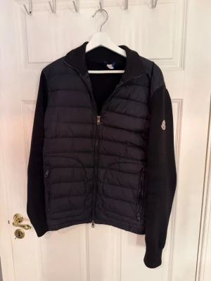 Svart Moncler cardigan  - Säljer en svart cardigan från Moncler med quiltad front och mjuka stickade ärmar. Jackan har två fickor med dragkedja och en liten Moncler-logga på ena ärmen. Perfekt för dig som vill ha en stilren och bekväm jacka.