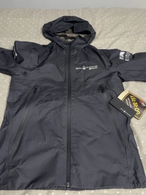 Svart vindjacka från Sail Racing - Snygg svart vindjacka från Sail Racing med huva och dragkedja framtill. Jackan är tillverkad i slitstark Gore-Tex och har två praktiska fickor med dragkedja. Perfekt för blåsiga dagar och outdoor-äventyr.