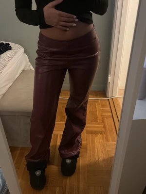 Vinröda skinnbyxor med straight fit - Säljer ett par vinröda skinnbyxor med rak passform och låg midja. Byxorna har en clean look utan synliga fickor eller detaljer och är tillverkade i ett glansigt skinnmaterial.