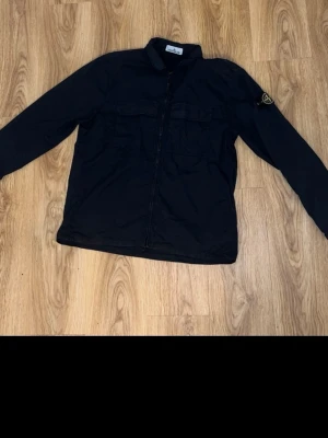 Svart overshirt från Stone Island - Säljer en svart overshirt från Stone Island med klassisk patch på ärmen. Jackan har dragkedja framtill, två bröstfickor och en clean, stilren look. Perfekt för dig som gillar streetwear och vill ha något enkelt men ändå fett.
