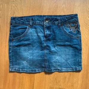 Blå jeanskjol med snygga detaljer  - Säljer en kort blå jeanskjol med snygga broderade detaljer och små nitar på fickorna fram och bak. Passar S/M. 