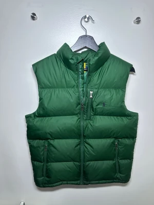 Grön dunväst från Polo Ralph Lauren - Säljer en grön dunväst från Polo Ralph Lauren med hög krage och två stora fickor med dragkedja. Västen har quiltad design och en diskret logga på bröstet. Storleken är 18-20 år men sitter som S i herrstorlek. 🟢✅