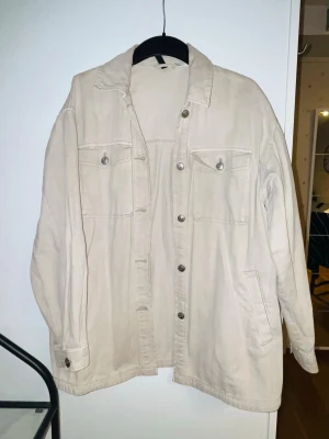 Beige overshirt i jeansmaterial - Säljer en beige overshirt i jeansmaterial med klassisk krage och knappar framtill. Två bröstfickor med lock och knapp samt sidofickor. Perfekt att slänga över en hoodie eller t-shirt för en chill look.