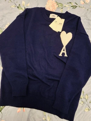 Marinblå stickad tröja från Ami - Säljer en marinblå stickad tröja från Ami Alexandre Mattiussi med ikoniskt vitt hjärta och A på bröstet. Tröjan har rund hals, ribbade muddar och är långärmad. Perfekt för dig som vill ha en stilren och trendig look.