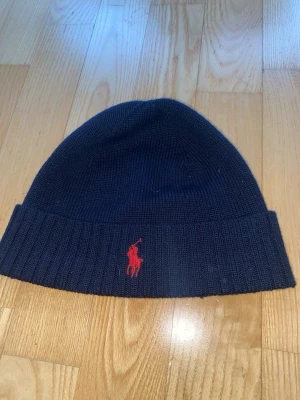 Marinblå mössa från Polo Ralph Lauren - Säljer en marinblå mössa från Polo Ralph Lauren med röd broderad logga framtill. Mössan är ribbstickad och tillverkad i 100% merinoull vilket gör den både mjuk och varm. Perfekt accessoar för kyliga dagar.
