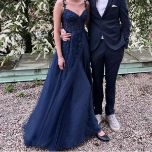 Marinblå balklänning med spetsdetaljer - Säljer en marinblå balklänning /festklänning med tunna axelband dekorerade med blommor. Klänningen har v-ringad urringning, spetsdetaljer på livet och ett luftigt, svepande kjolparti i tyll. Perfekt för bal eller student! Sitter snyggt och ger en elegant siluett.