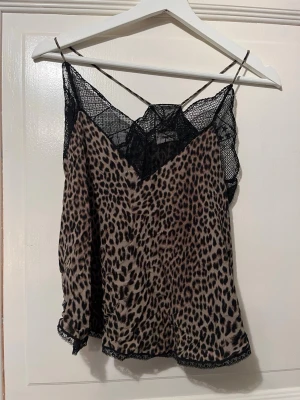 Leopardmönstrad linne Zadig & Voltaire - Snygg leopardmönstrad linnetopp från Zadig & Voltaire med tunna axelband och svart spetsdetalj upptill och nedtill. Toppen har en v-ringad skärning och är gjord i ett glansigt, mjukt material som känns lyxigt.
