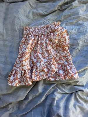 Blommig volangkjol i aprikos och vitt - Supersöt kort kjol med blommigt mönster i aprikos och vitt. Kjolen har volangdetaljer och en smockad midja som ger en skön passform. Perfekt för dig som gillar en feminin och trendig stil.