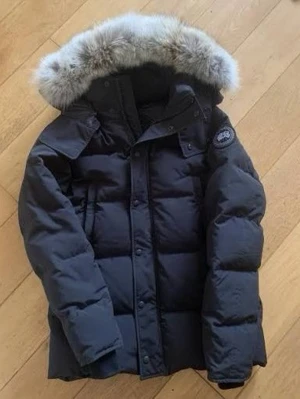 Svart dunjacka från Canada Goose Label - Säljer en svart dunjacka från Canada Goose Label med fluffig päls på huvan och klassisk patch på ärmen. Jackan har quiltad design, långa ärmar och stängs med både dragkedja och knappar. Perfekt för kalla vinterdagar och riktigt snygg till alla outfits.