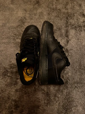 Nike Air Force 1 svart/gul sneakers - Säljer ett par svarta Nike Air Force 1 sneakers med gula detaljer och logga. Skorna har klassisk låg siluett, snörning och kombinerar skinn och textil för en clean look. Swoosh-loggan har gul kant och innersulan är gul för extra kontrast.